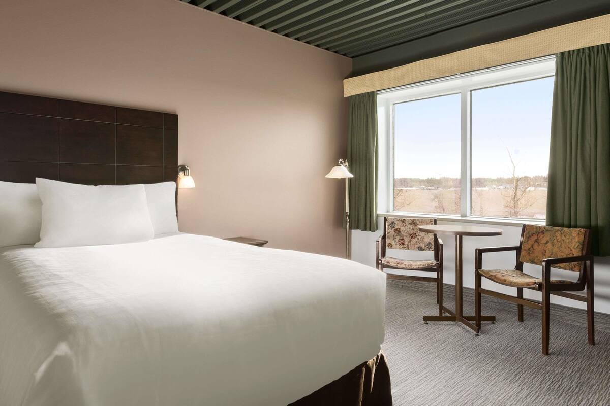 Ein Hotelzimmer mit einem großen Bett, zwei Sesseln, einem kleinen Tisch und einem Fenster mit Vorhängen.