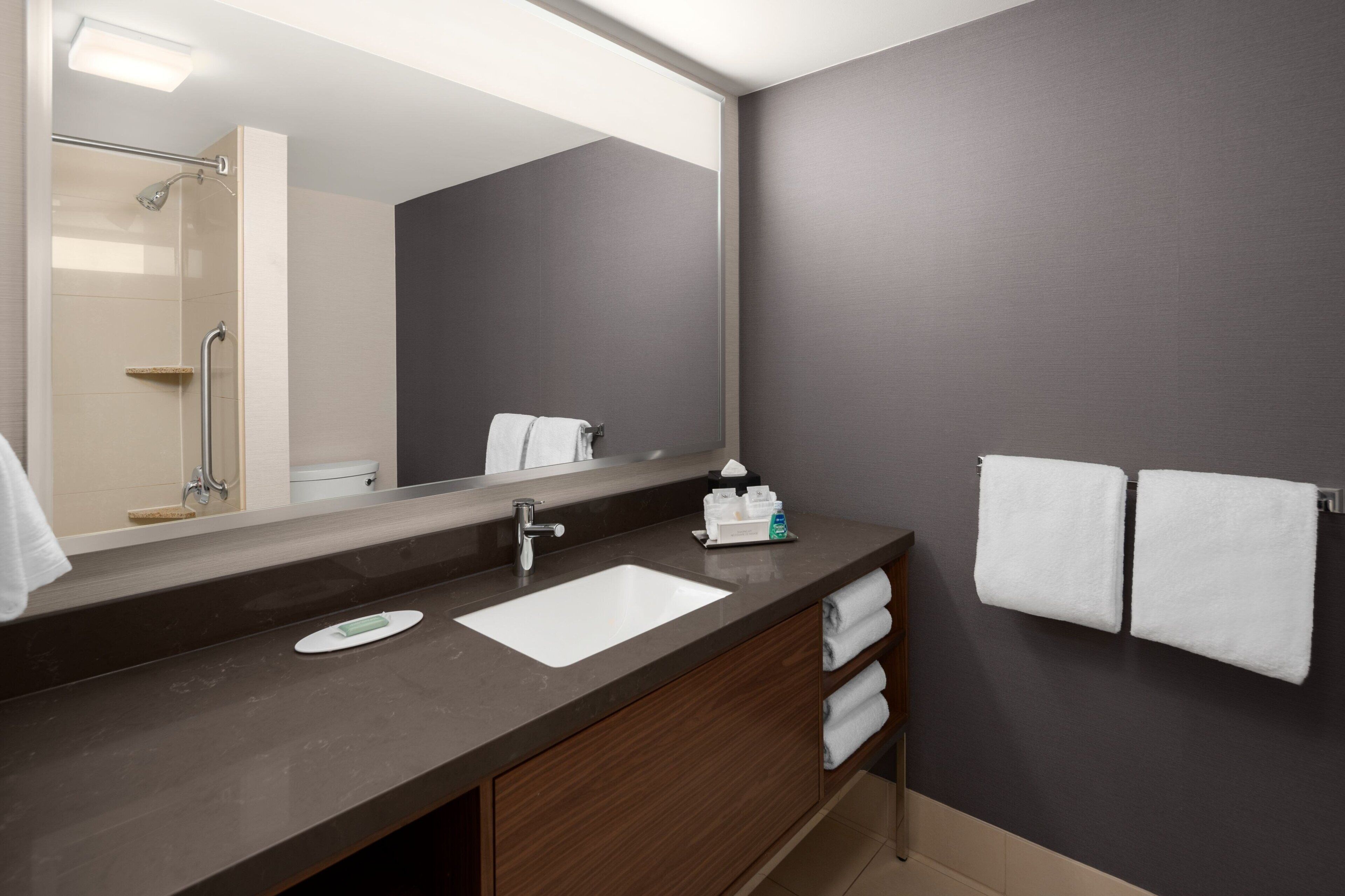 A modern bathroom with a large mirror, a sink, and a shower. There are towels and toiletries on the counter.