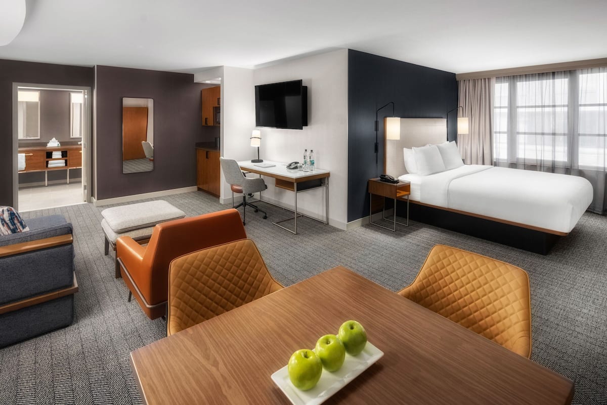 A hotel room with a bed, desk, chair, and a table with apples.