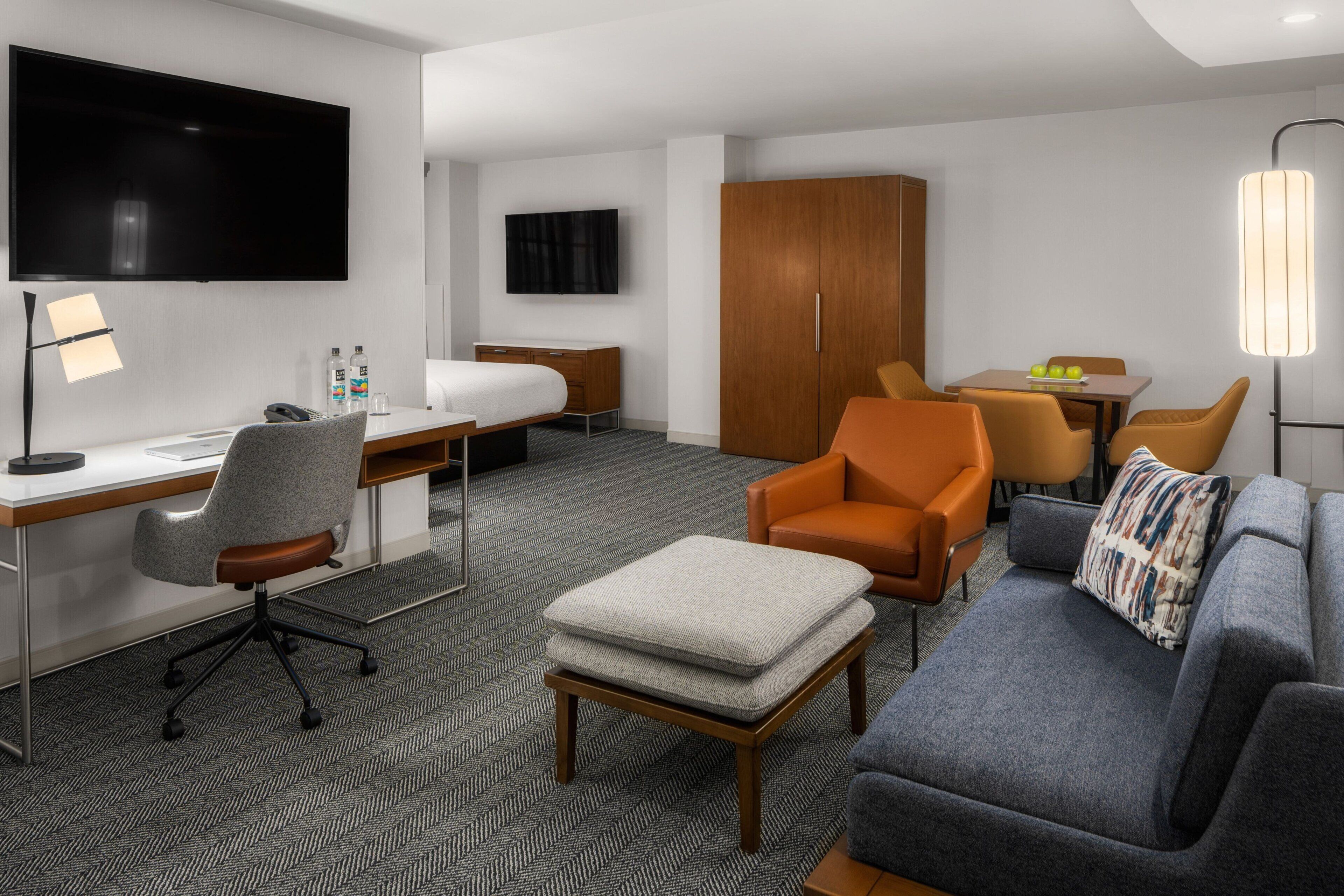 Une chambre d’hôtel équipée d’une télévision à écran plat, d’un bureau avec une chaise, d’un canapé, d’un pouf, d’une table à manger avec des chaises et d’une armoire.