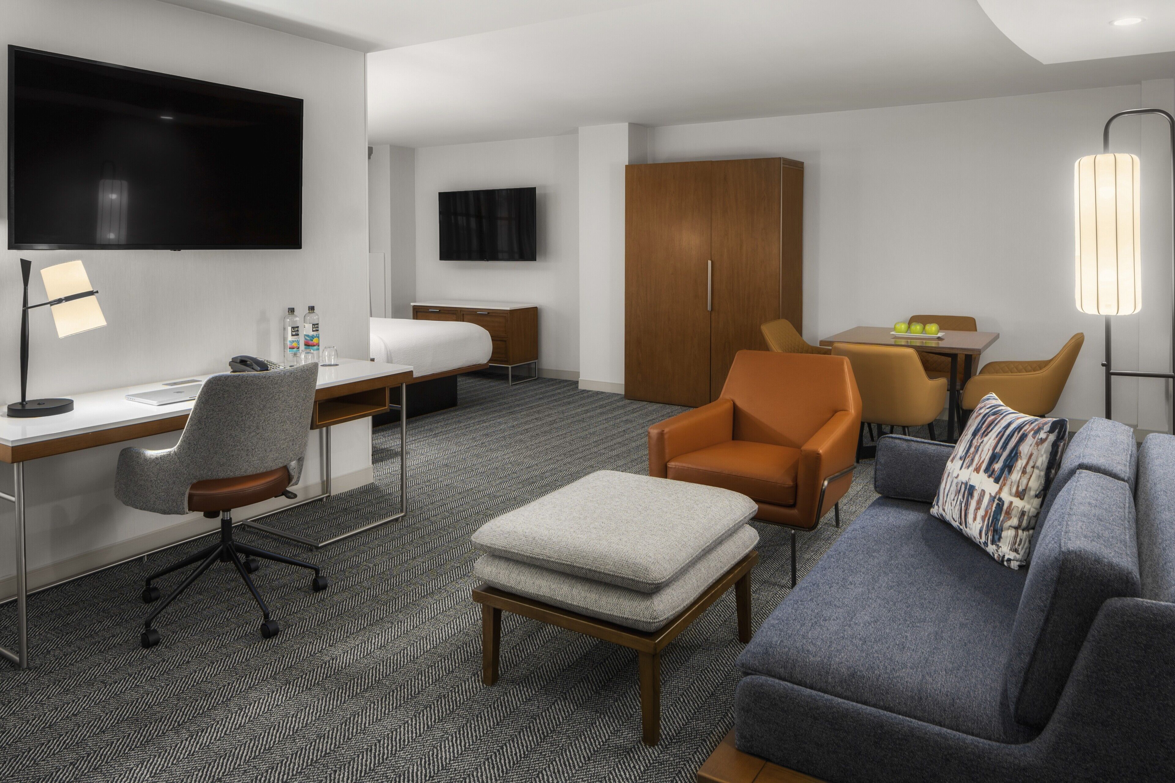 A hotel room with a desk, office chair, sofa, ottoman, TV, wardrobe, and a dining area with chairs and table.