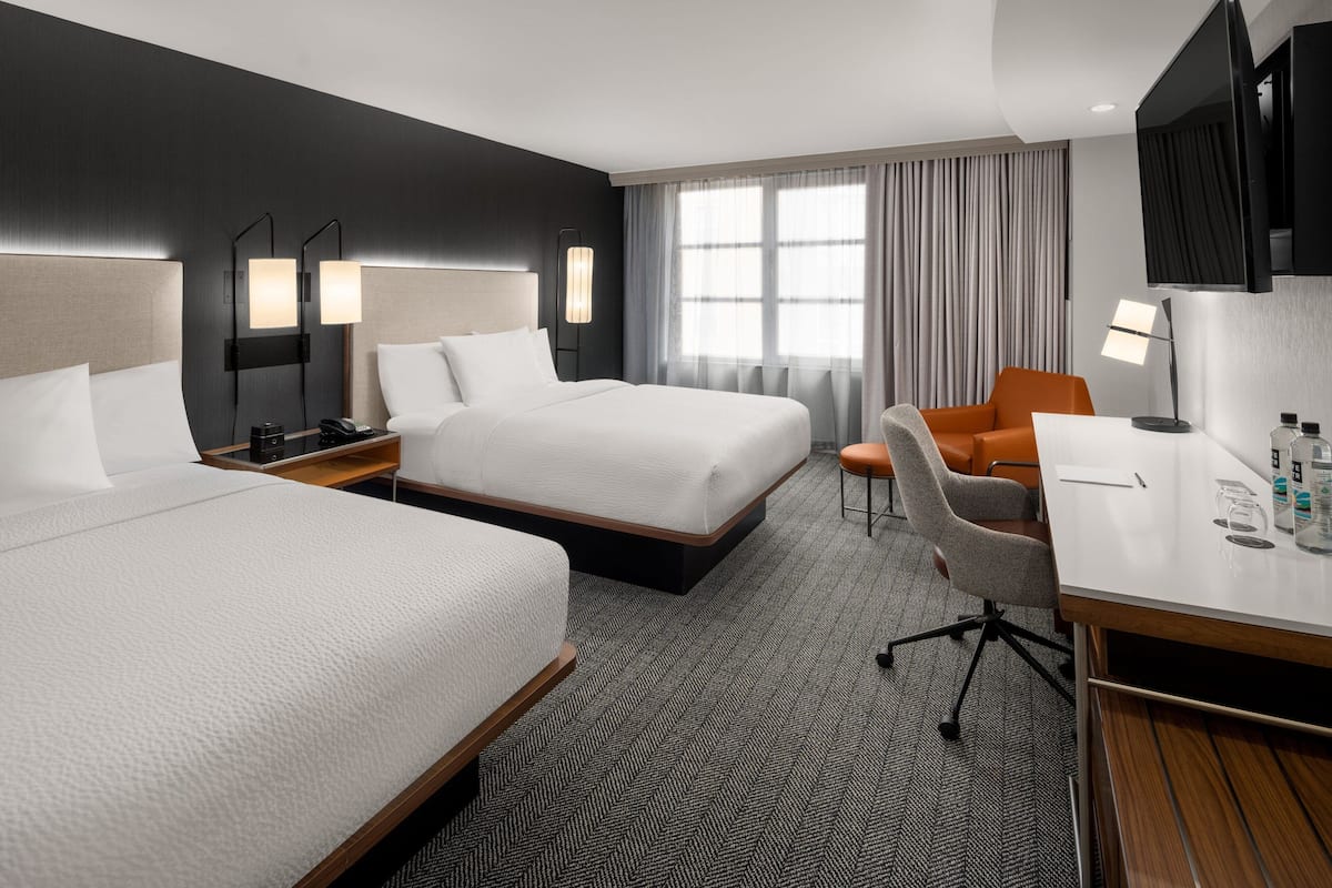 A hotel room with two beds, a desk, an orange chair, and a TV mounted on the wall.