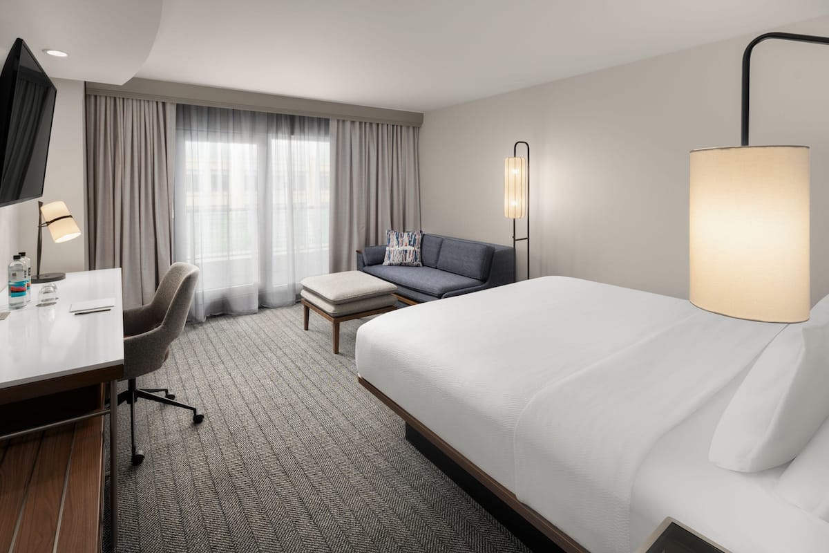 A hotel room with a bed, a desk, a chair, a sofa, a lamp, and a window with curtains.