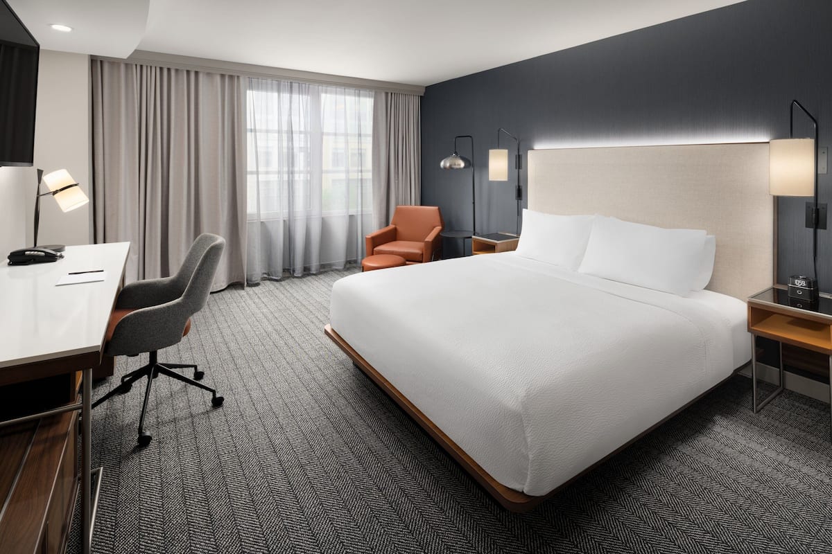 A hotel room with a large bed, a desk with a chair, a small table, and a lamp.