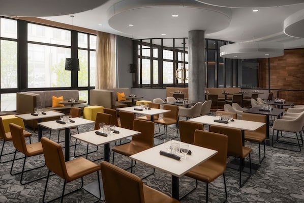 Un espace de restauration moderne dans un hôtel, avec de nombreuses tables et chaises, de grandes fenêtres et un coin salon central agrémenté d’un canapé et de coussins.