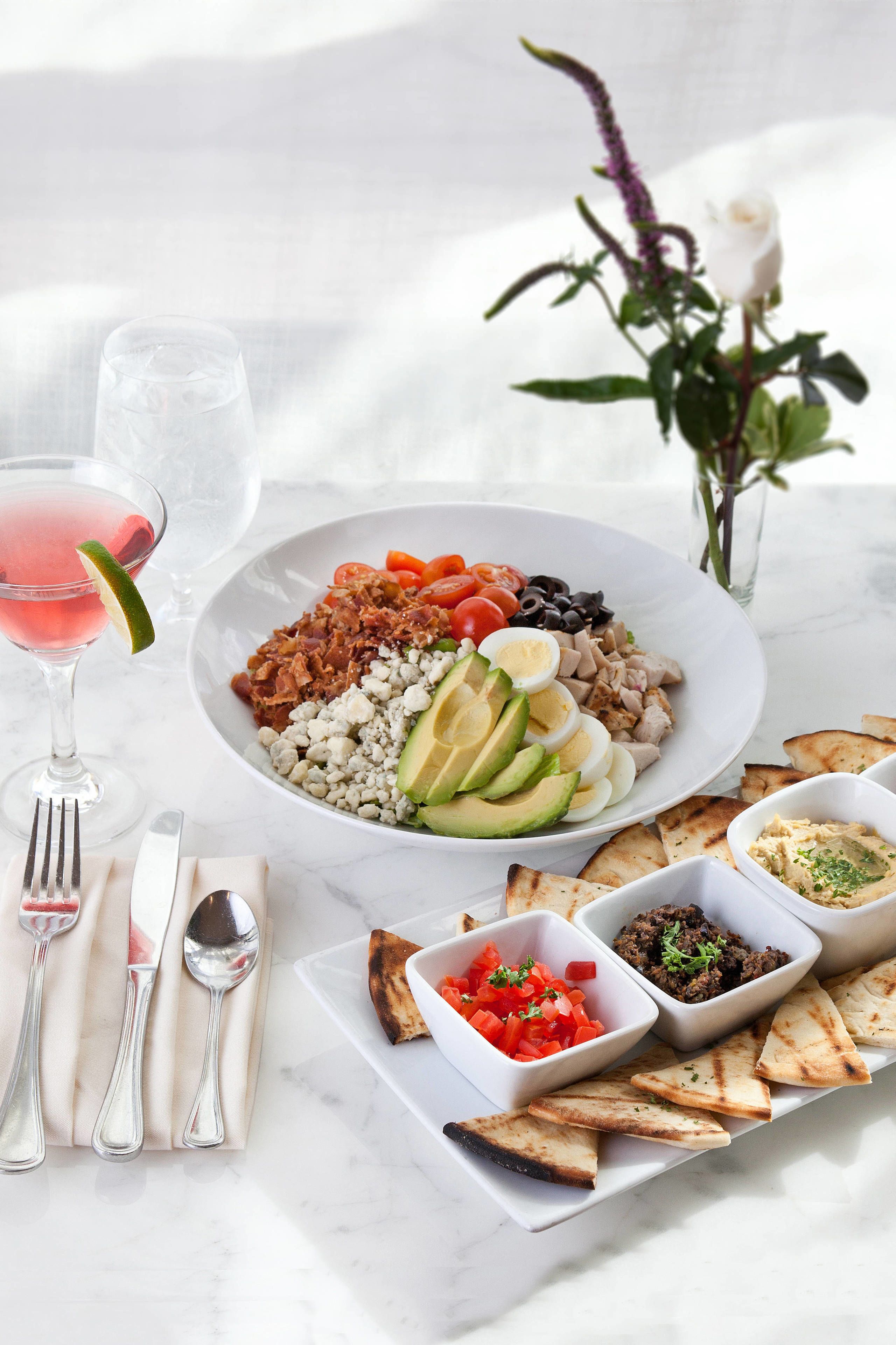 A white plate with a salad, a glass of pink drink, and a vase with a white rose.