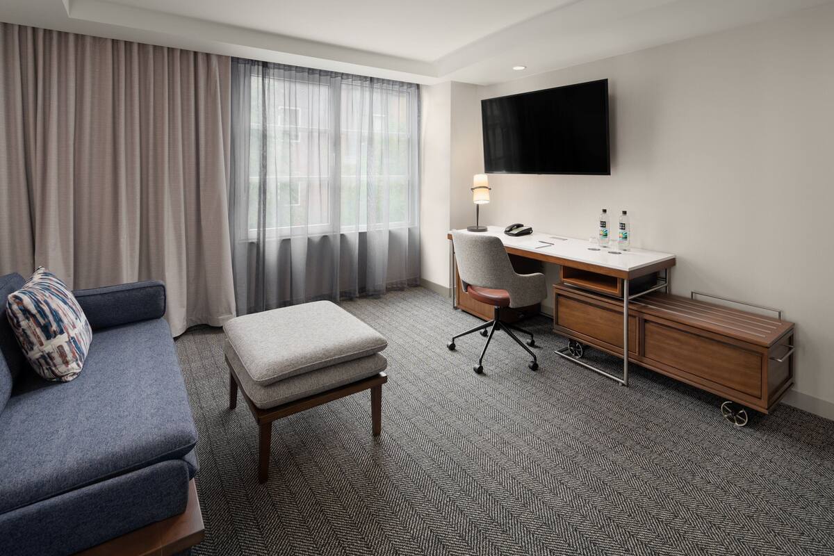 A hotel room with a sofa, a desk, a chair, a TV, and a minibar.