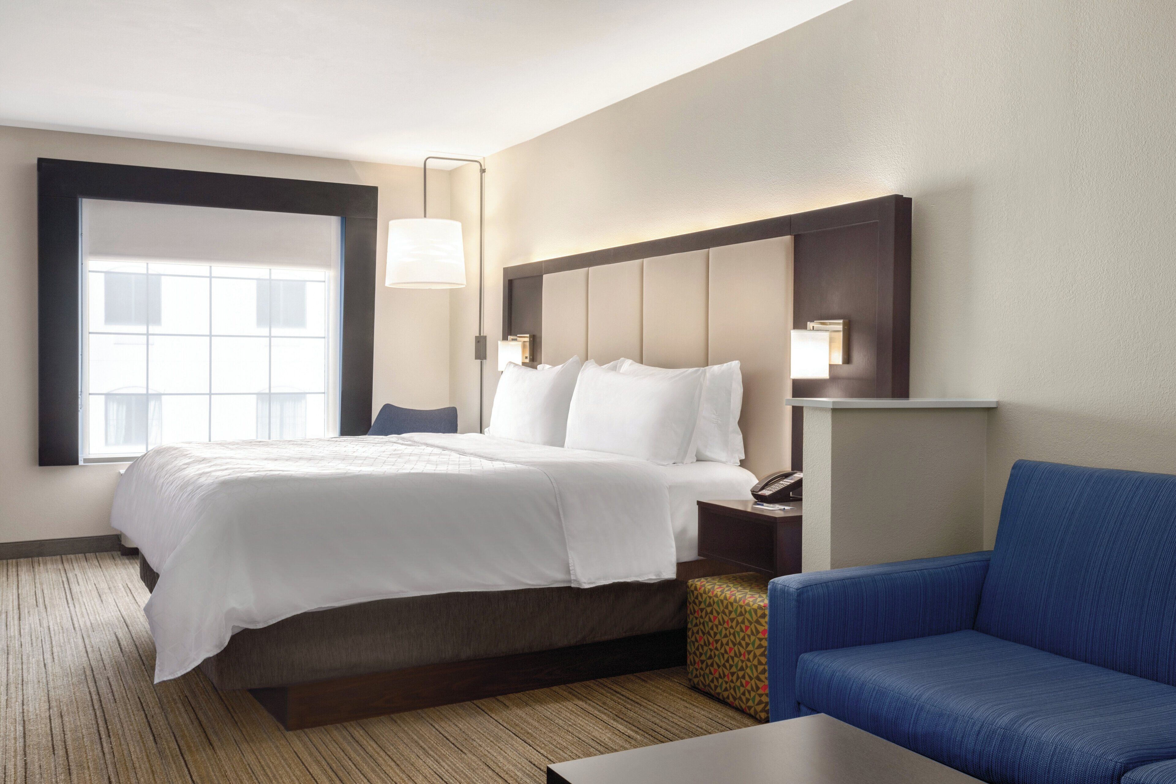 A hotel room with a large bed, a bedside table with a lamp, a blue sofa, and a window.