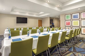 Un salón de conferencias con mesas, sillas, botellas de agua y una pantalla de televisión.