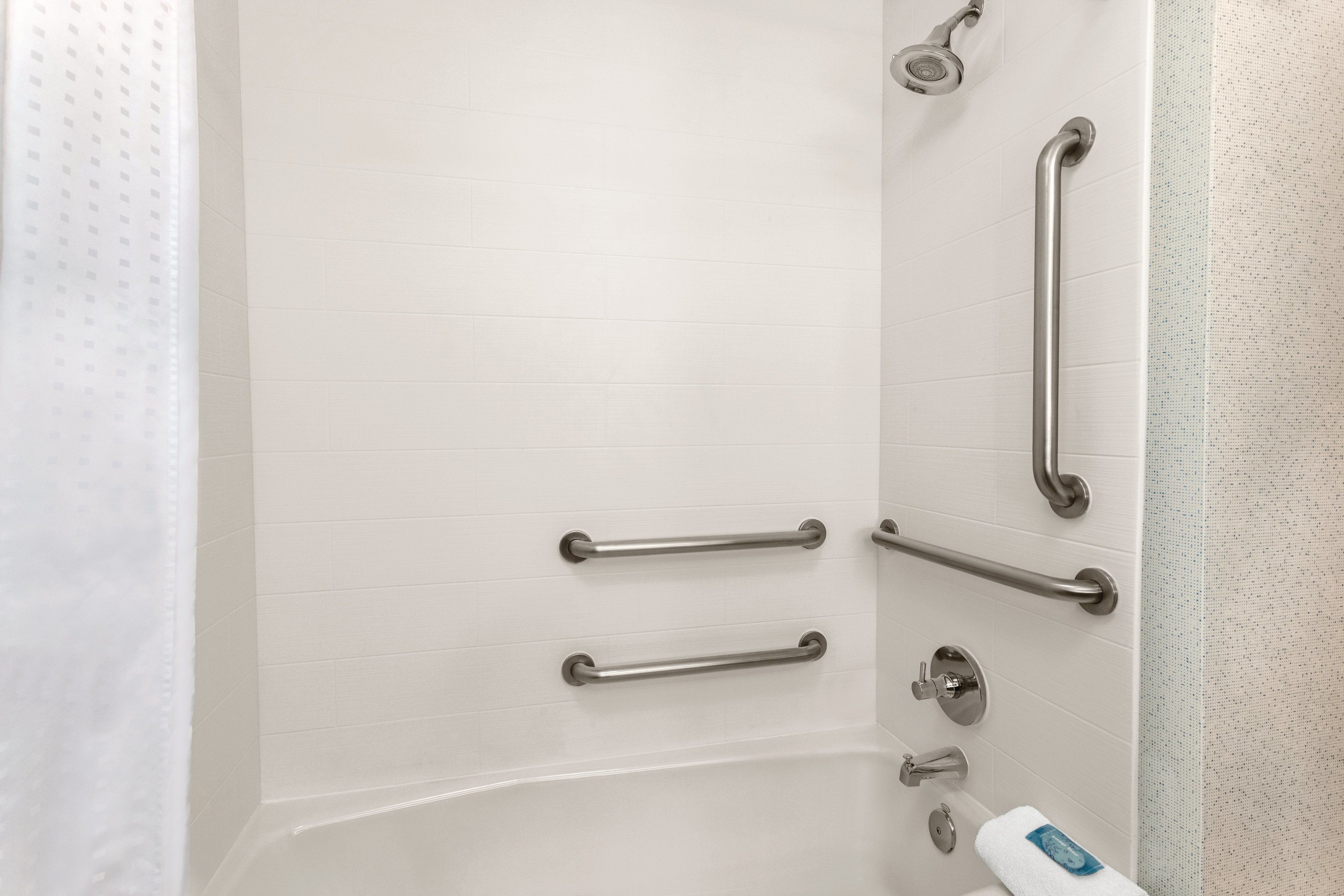 Une salle de bain avec des murs recouverts de carreaux blancs, une baignoire et plusieurs barres d’appui installées pour plus de sécurité.