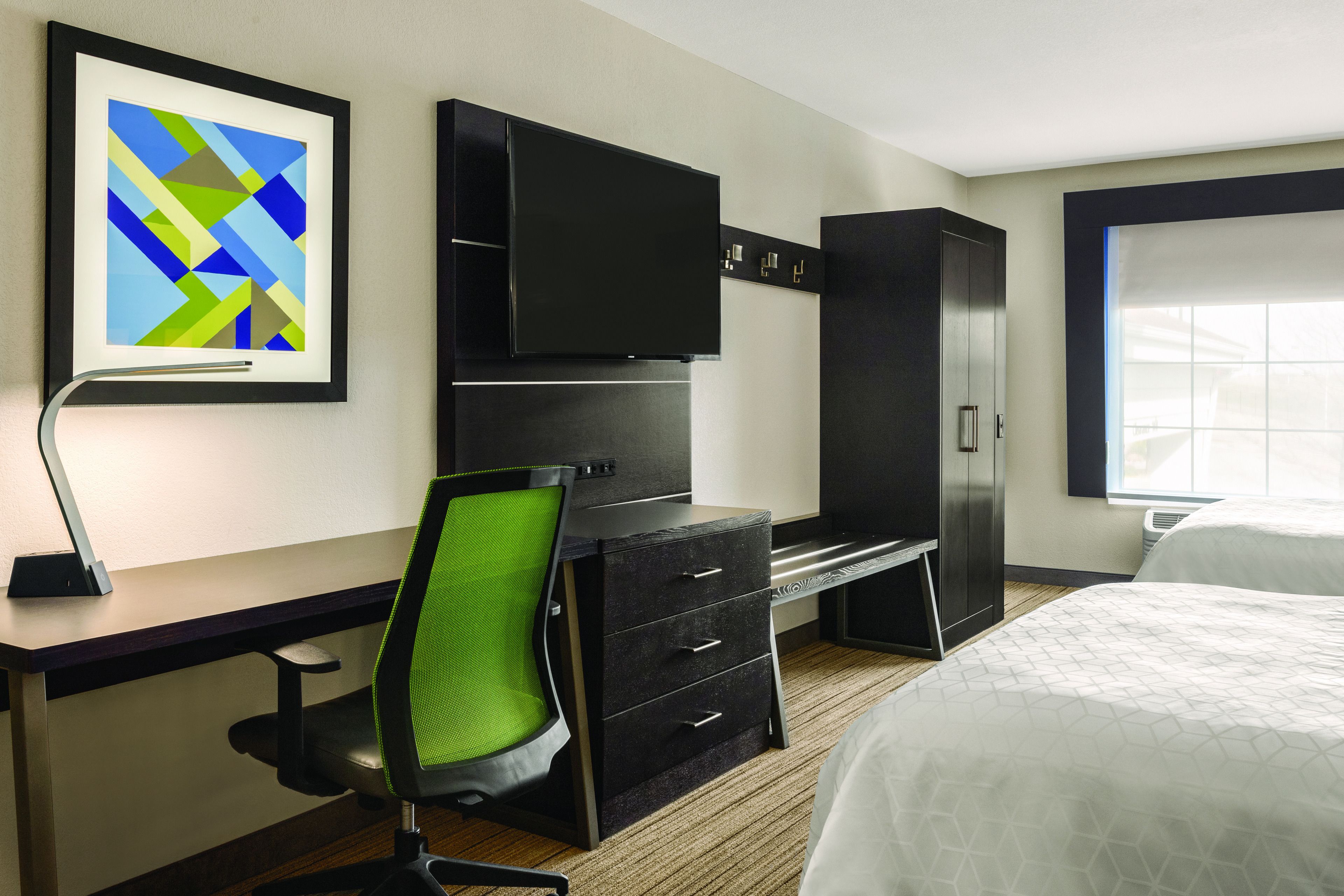 A hotel room with a desk, chair, wardrobe, bed, artwork, and a window.