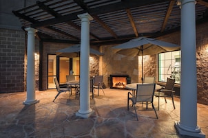 Un patio con suelo de piedra, mobiliario exterior, chimenea y una zona cubierta con columnas.