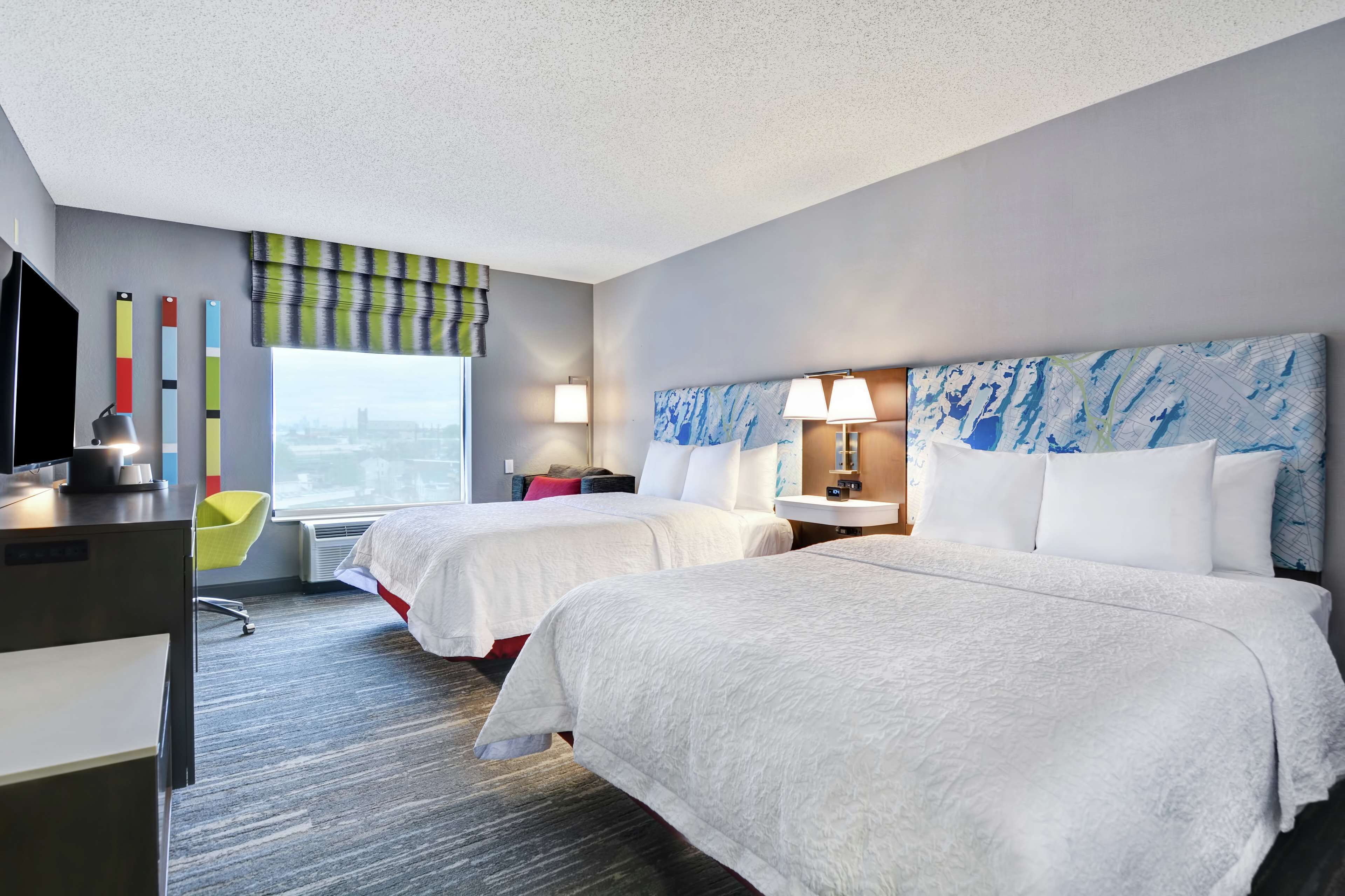 A hotel room with two beds, a desk with a computer, a chair, and a window with a view of the city.