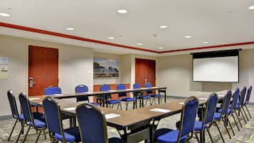 A conference room with a long table, blue chairs, a projector screen, and a carpeted floor.