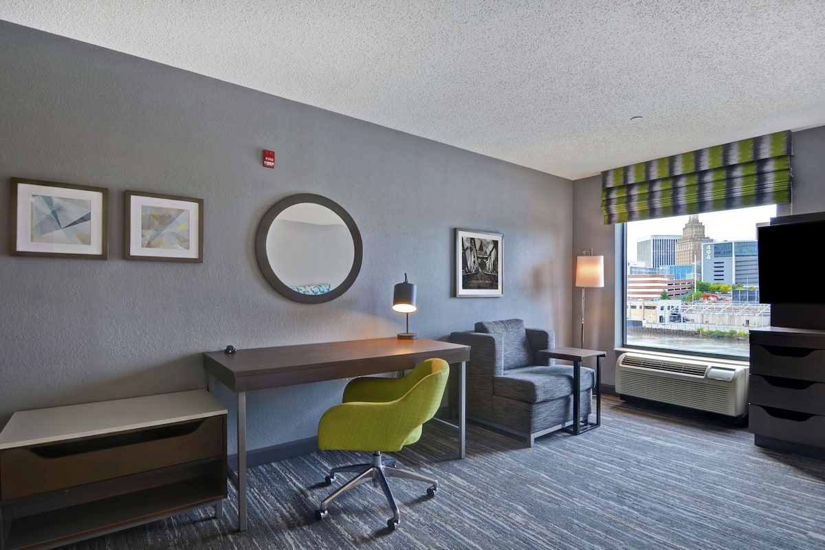 Habitación de hotel con escritorio, una silla verde, sofá, espejo redondo y ventana con vista a la ciudad.