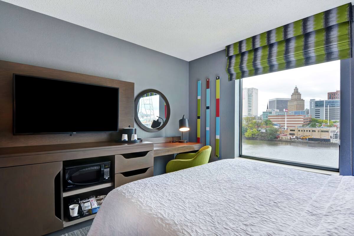 Habitación de hotel con cama, televisión, escritorio con silla, microondas y vistas a un paisaje urbano a través de un ventanal grande.