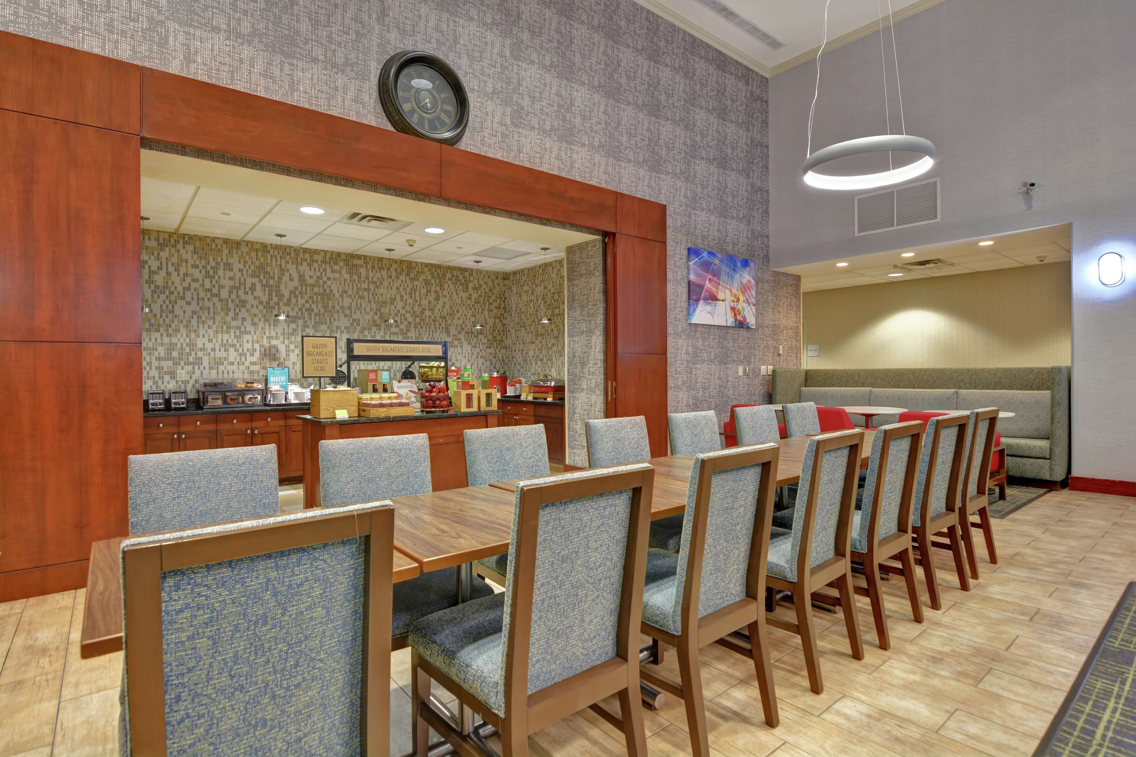 A hotel dining area with a long wooden table, multiple chairs, a bar area, and a seating section with a sofa.