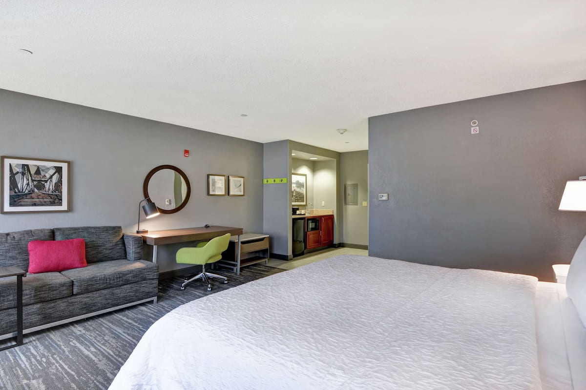 Habitación de hotel con una cama grande, un sofá gris con un cojín rojo, un escritorio con una silla y una pequeña cocina americana al fondo.