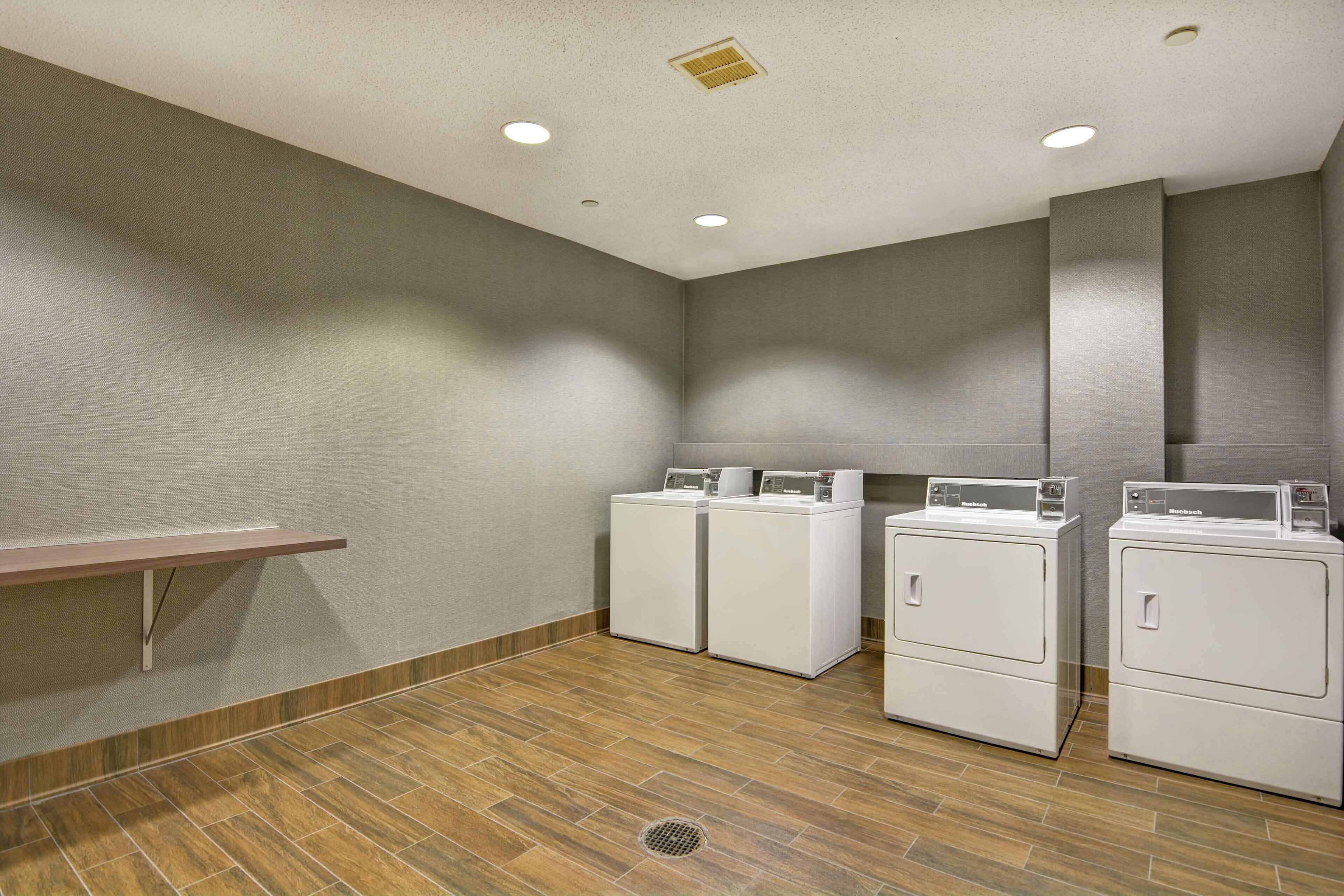 A laundry room with three stacked washers and dryers, a wooden bench, and a vent on the ceiling.