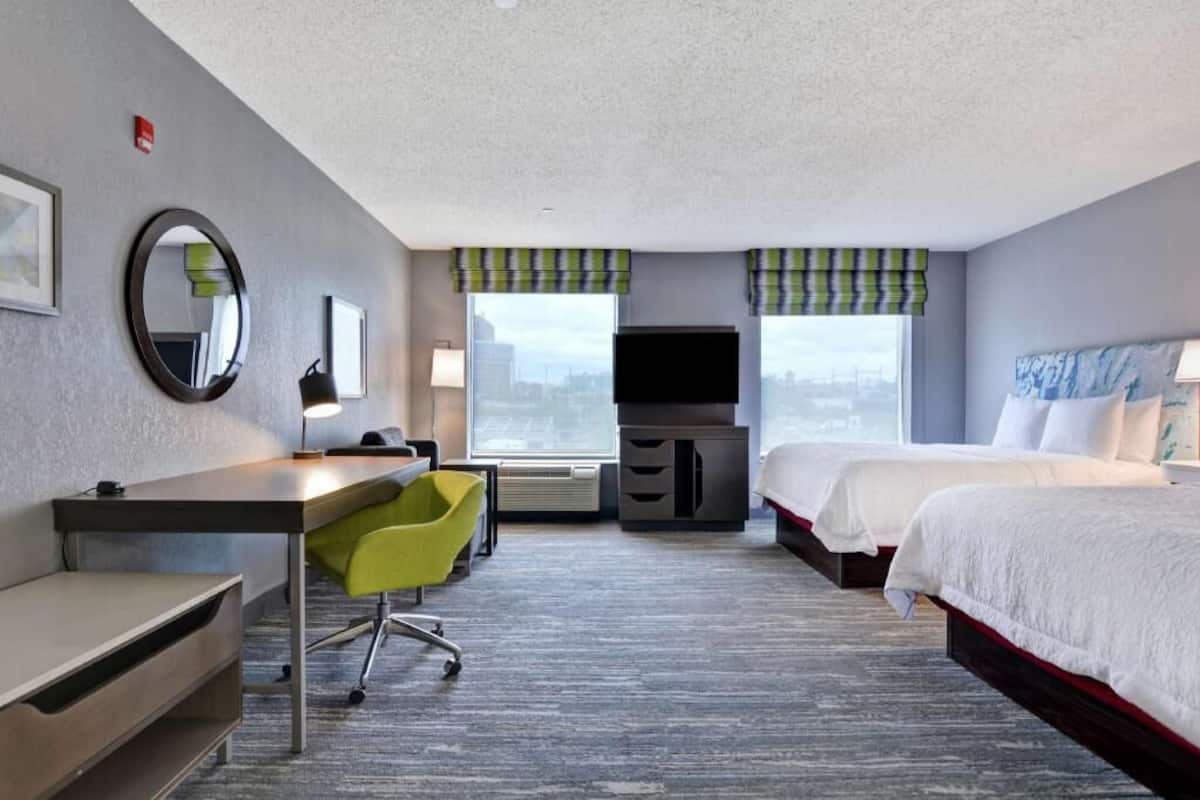 Habitación de hotel con escritorio, silla, cama, televisor y ventanas amplias.