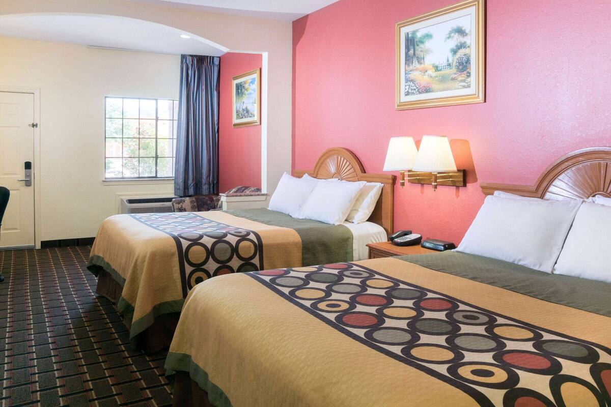 A hotel room with two beds, a window with curtains, a door, and a wall-mounted lamp.