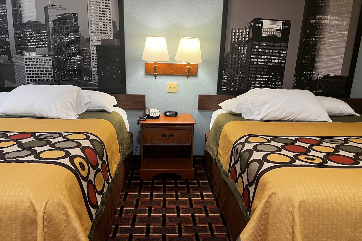 A hotel room with two beds, a nightstand with a phone and lamp, and a cityscape view.