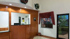 Un área de recepción con mostrador de madera, un tablón de anuncios, una ventana con vista a palmeras y una puerta con un cartel.