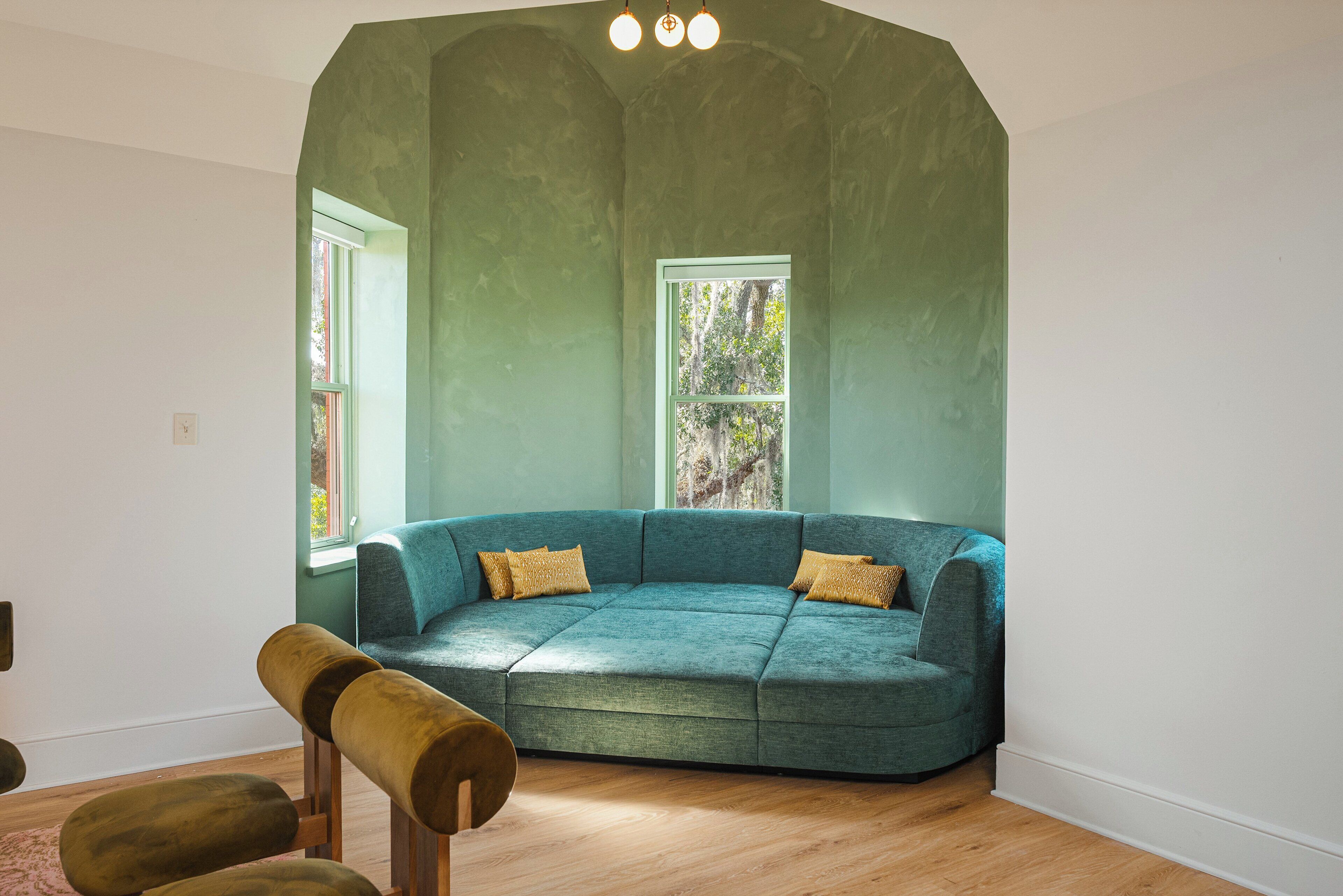 Una habitación con un sofá seccional color turquesa, dos ventanas, un banco de madera y una pared blanca.
