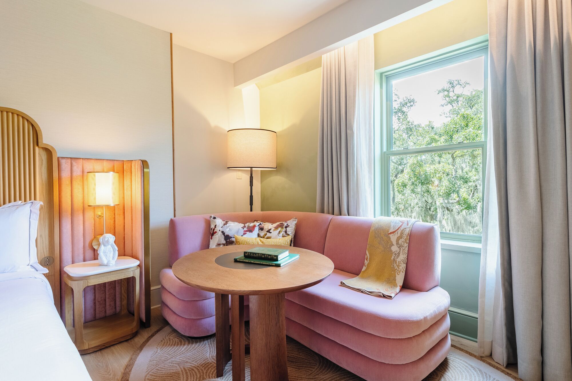 A hotel room with a pink sofa, a round wooden table, a lamp, and a view of trees through a window.