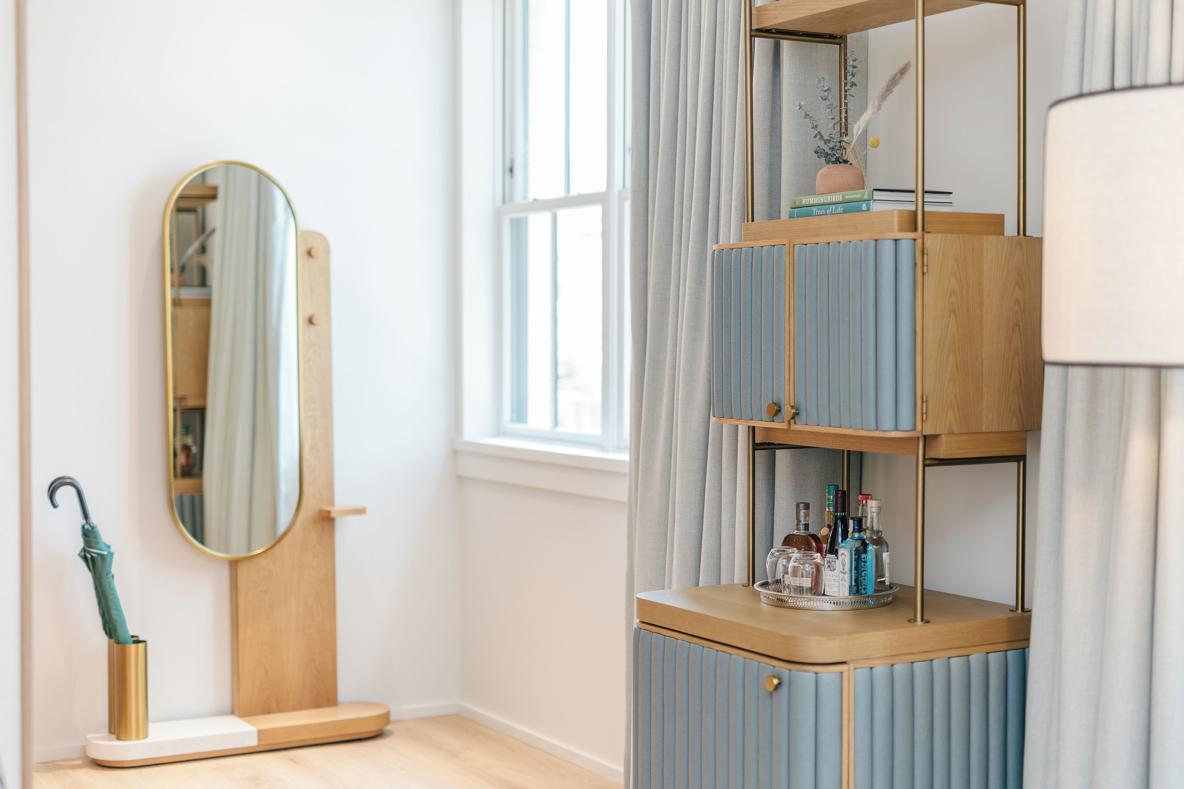 Una habitación con un tocador de madera, un espejo, un estante con libros y objetos decorativos, y una ventana con cortinas.