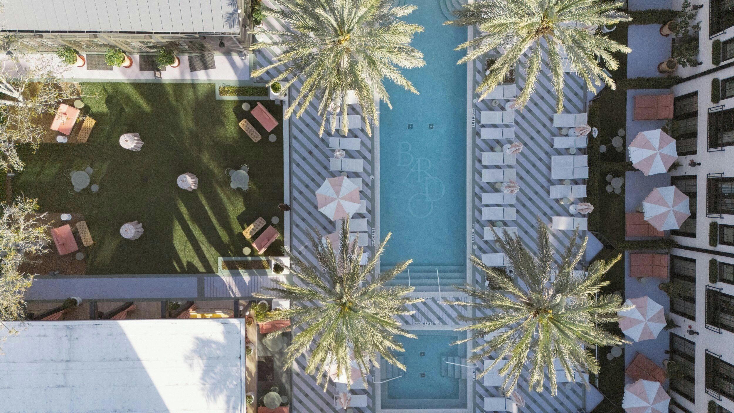 An aerial view of a hotel with a pool surrounded by lounge chairs and umbrellas, palm trees, and a rooftop bar area.