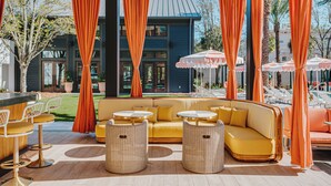 Een overdekt terras met gele banken en rieten tafels, oranje gordijnen en parasols.
