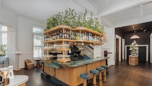 Una zona de bar con mostrador de madera, varias estanterías llenas de botellas y una planta colgante. Se ven taburetes de bar y una escalera de fondo.