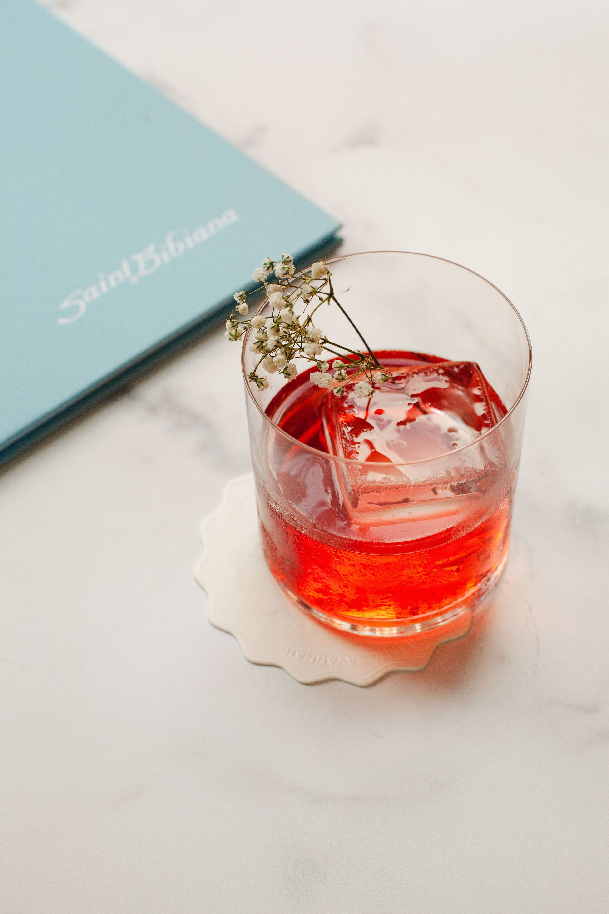 A glass of red beverage with a flower garnish on a white coaster, placed on a marble surface with a blue book in the background.