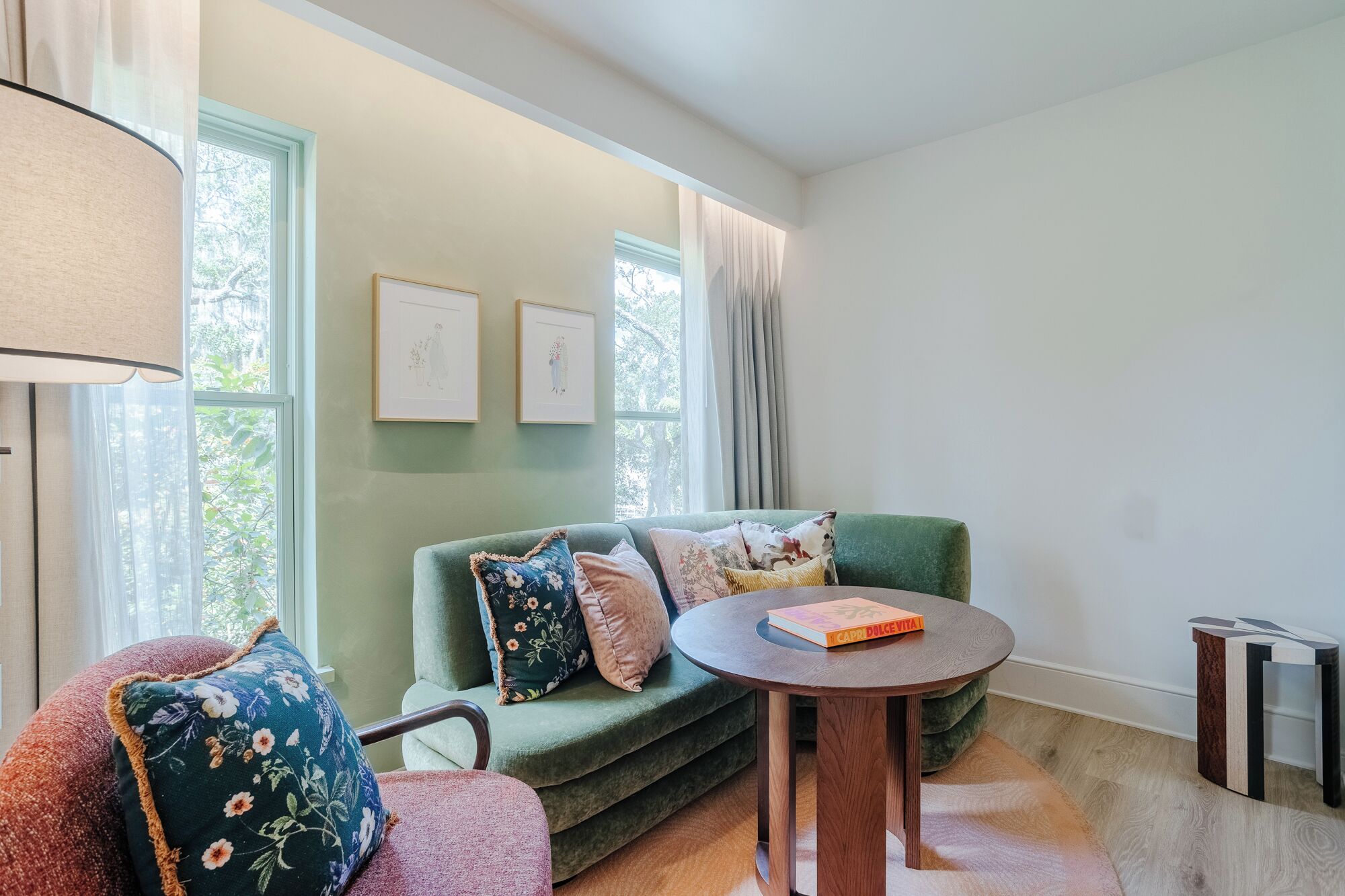Uma sala de estar aconchegante com um sofá verde, uma mesa de centro redonda de madeira e uma luminária. Há duas fotos emolduradas na parede e uma janela com cortinas.