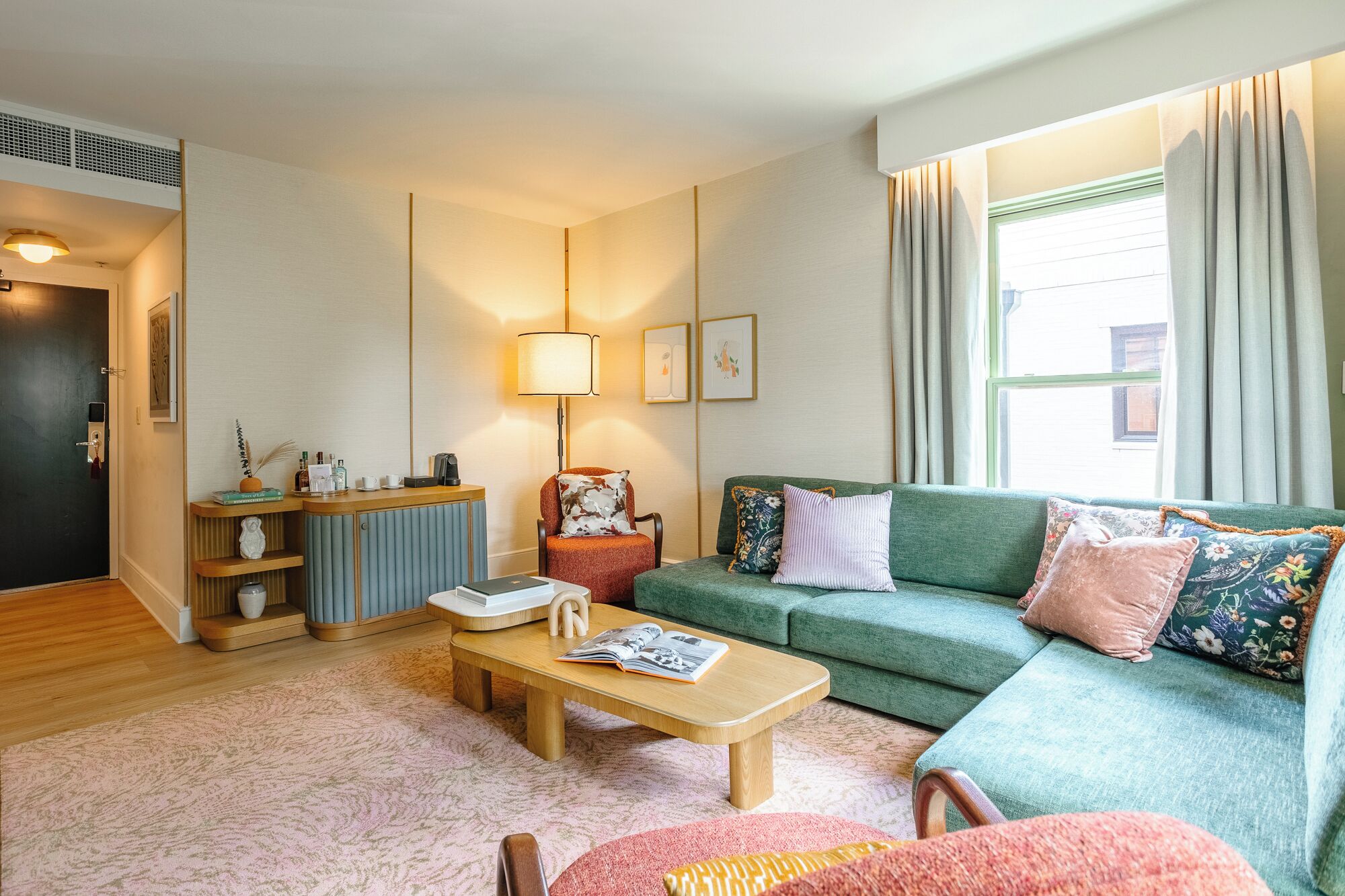 A cozy living room with a green sofa, a wooden coffee table, and a red armchair. There is a small side table with a lamp and decorative items. A window with curtains allows natural light into the room.