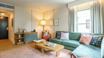 A cozy living room with a green sofa, a wooden coffee table, and a red armchair. There is a small side table with a lamp and decorative items. A window with curtains allows natural light into the room.