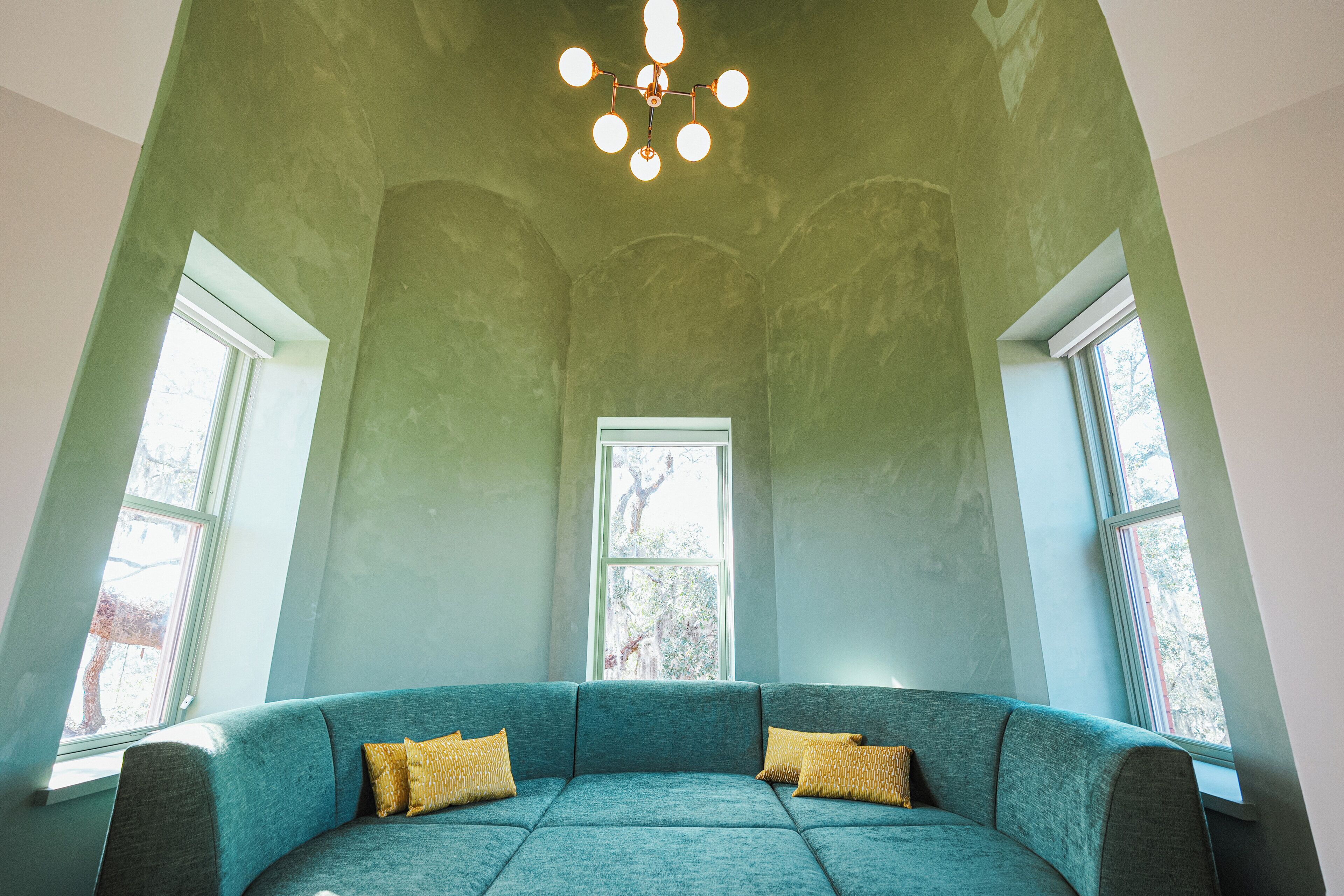 A room with a teal sofa, two windows, and a ceiling light fixture.