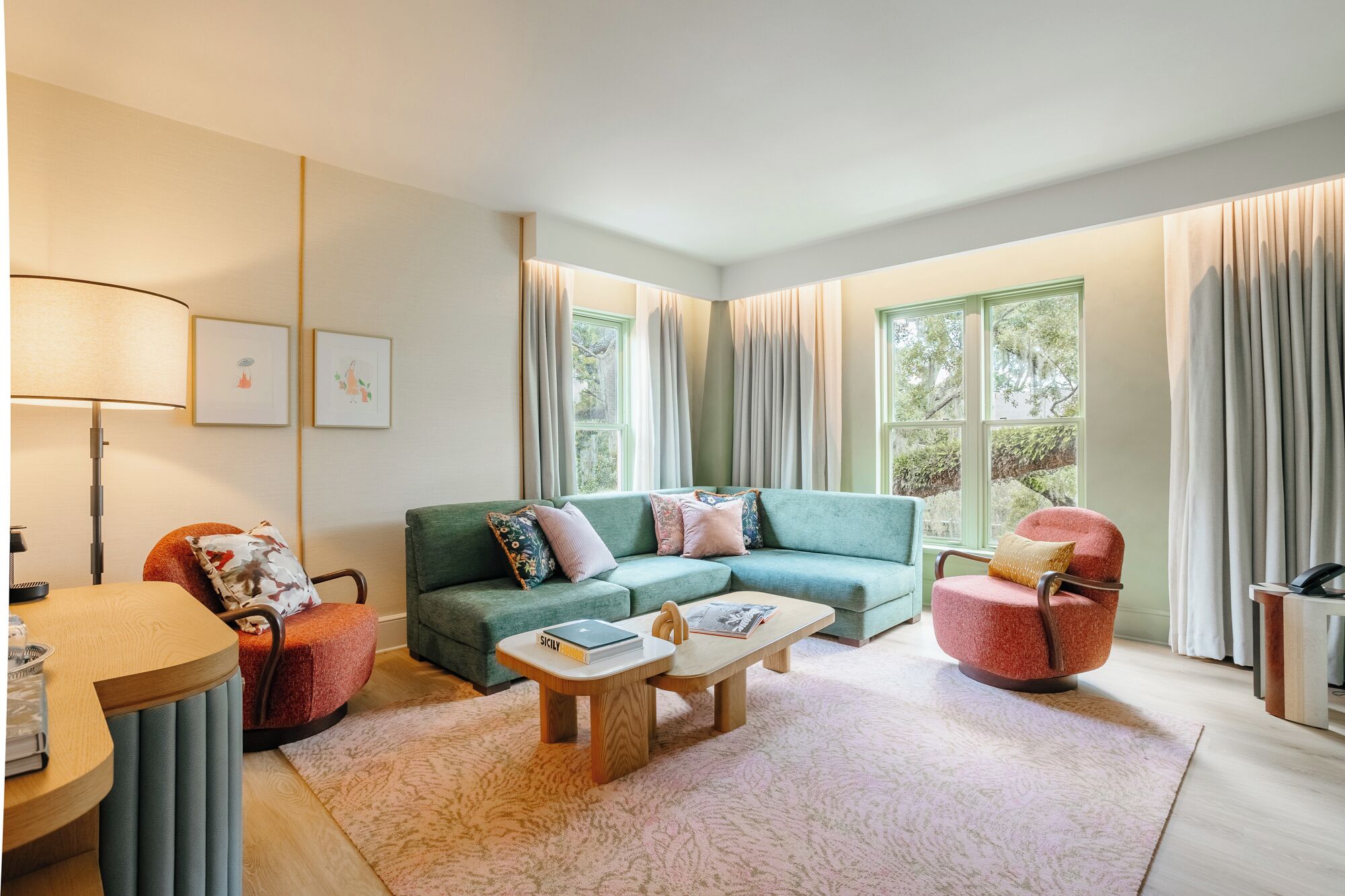 A modern living room with a green sofa, a red armchair, a wooden coffee table, and a lamp. There are two windows with curtains, and a framed picture on the wall.