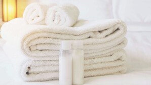 A stack of white towels and a bottle on a bed. - M star Hotel Searcy (Searcy)