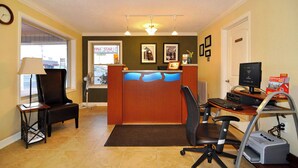 A reception area with a desk, computer, printer, and a chair. There is a window with a view of the outside, a clock on the wall, and framed pictures on the wall. - M star Hotel Searcy (Searcy)