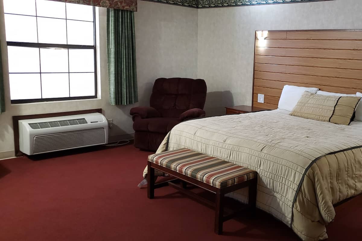 A hotel room with a large bed, a chair, a bench, a bedside table, a nightstand, and a window with curtains.