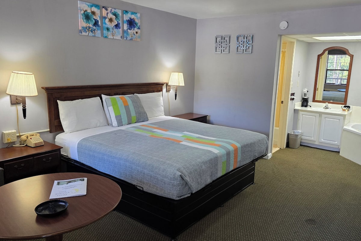 A hotel room with a bed, bedside lamps, a table with a chair, a small desk, a mirror, a sink, and a toilet.