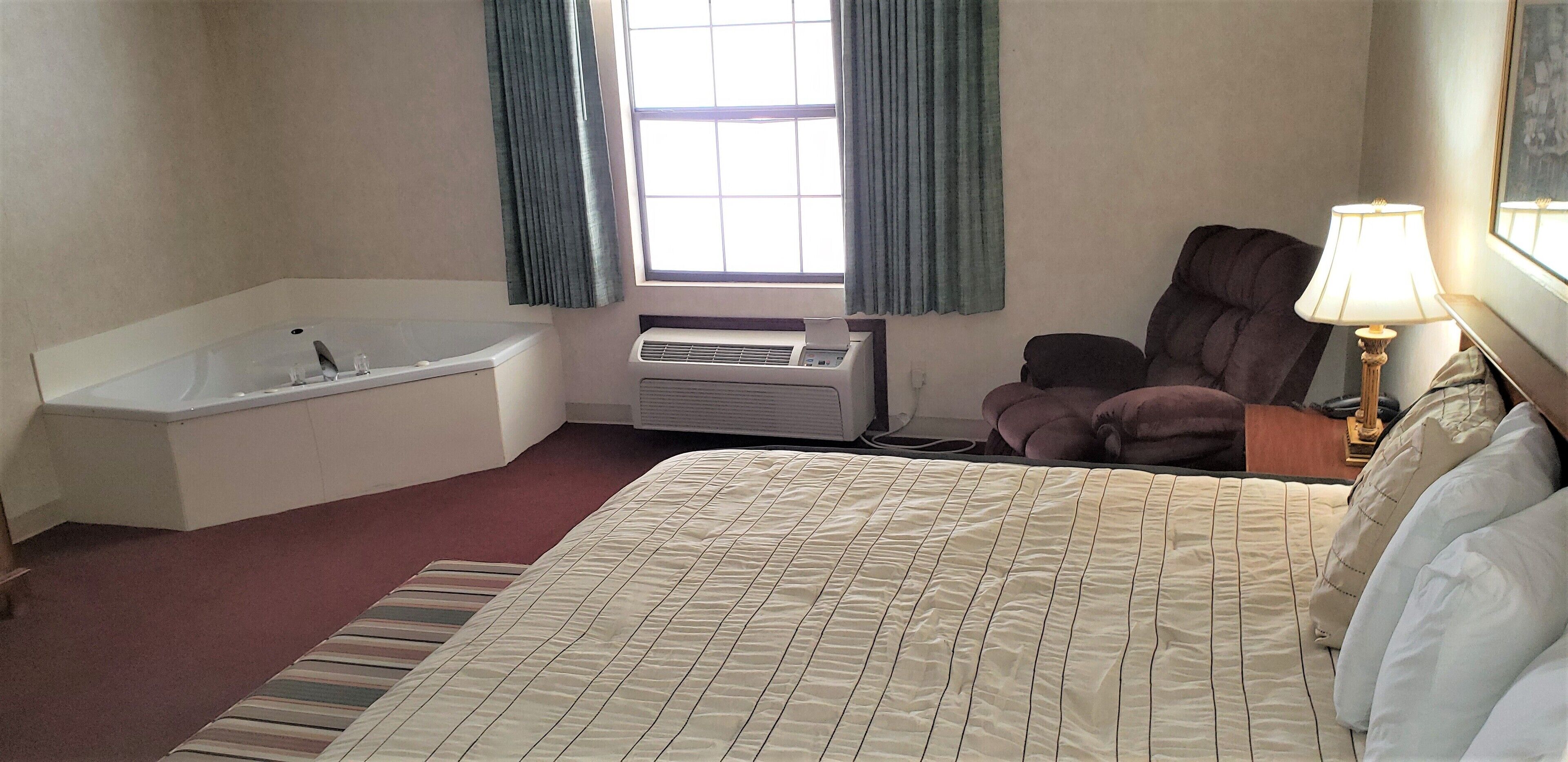 A hotel room with a large bed, a bathtub, a chair, a desk, a lamp, and a window with curtains.