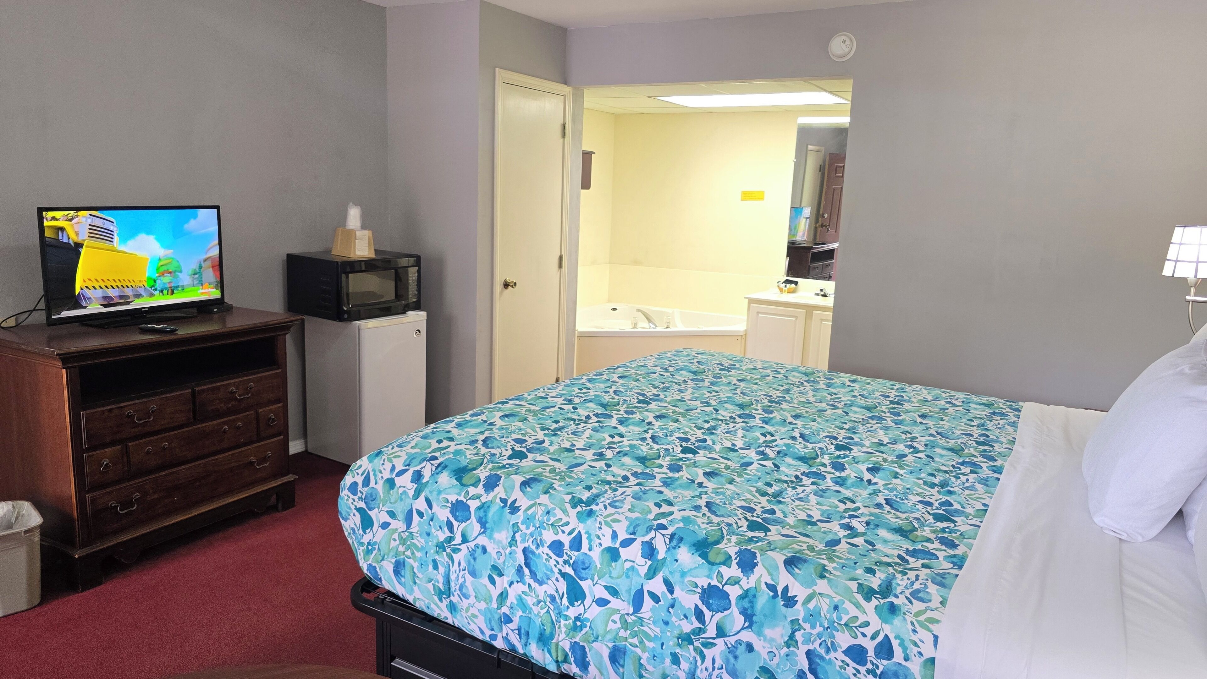 Habitación de hotel con cama, televisor sobre un aparador, refrigerador pequeño, baño visible a través de una puerta abierta y un cubo de basura.