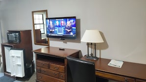 Habitación de hotel con escritorio de madera, televisor mostrando un noticiero, microondas, refrigerador, lámpara y espejo.