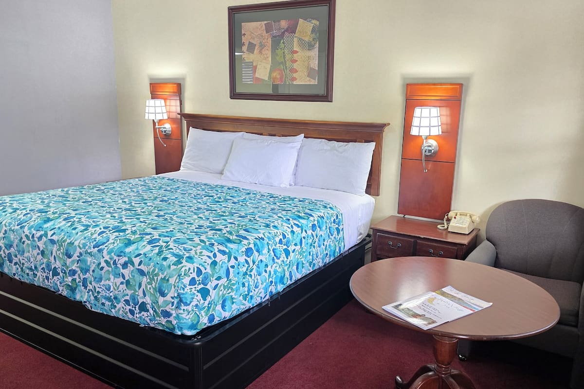 A hotel room with a bed, bedside lamps, a chair, a small table, and a framed artwork on the wall.