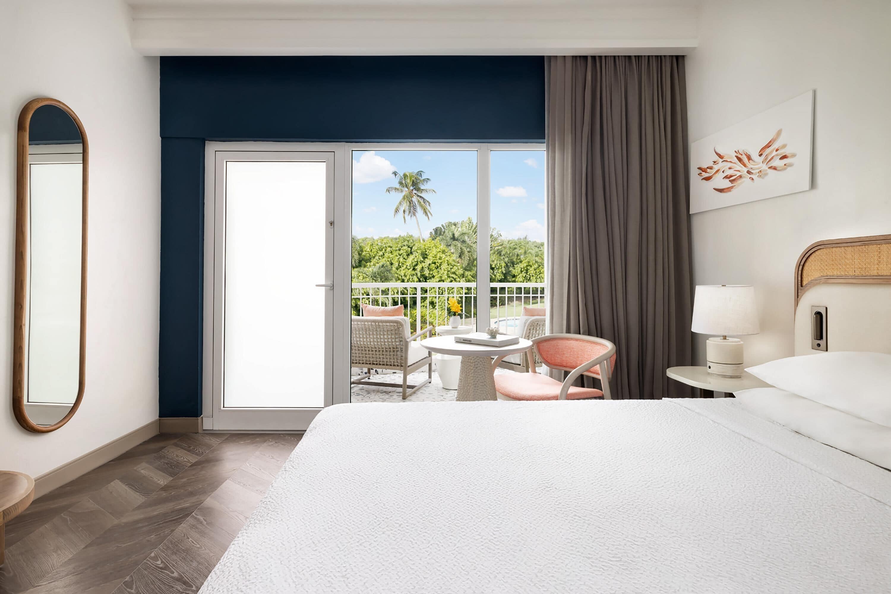 Habitación de hotel con cama, una mesa redonda, dos sillas, una mesita auxiliar, una lámpara, un espejo y un balcón con vistas a la vegetación.