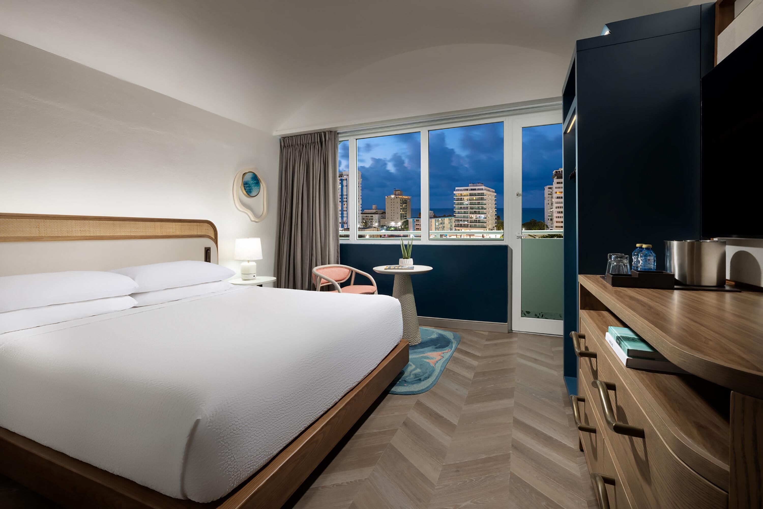 Habitación de hotel con cama, una mesita, una silla, televisor y vista nocturna de la ciudad.