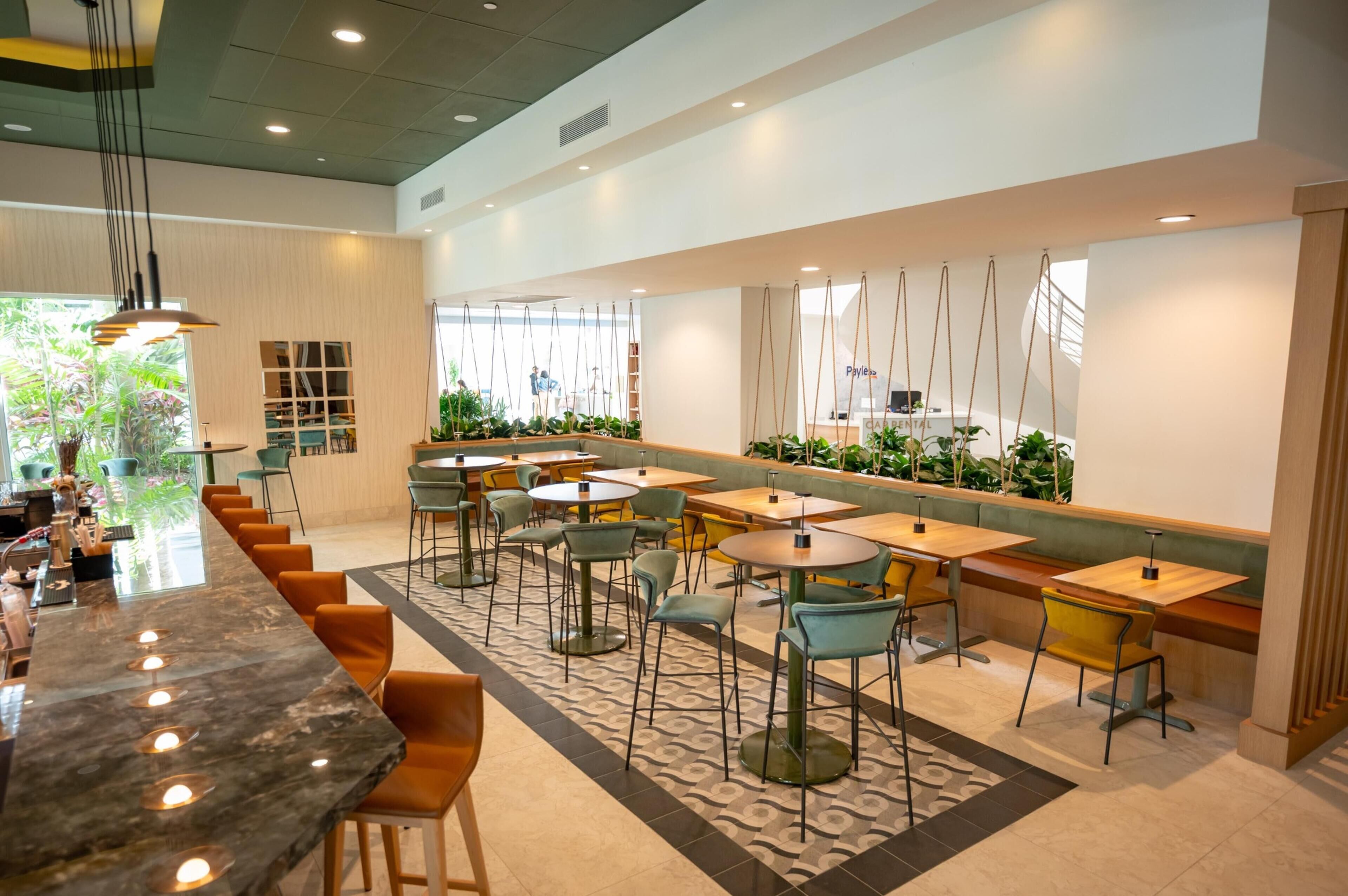 Un café moderno con una barra larga, varias zonas de estar y plantas de interior.
